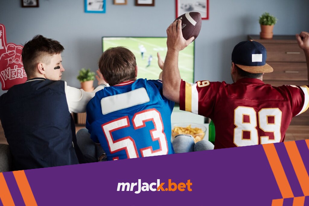 3 homens vistos de costas, vestindo camisas de times de futebol americano, assistindo ao jogo pela teve.