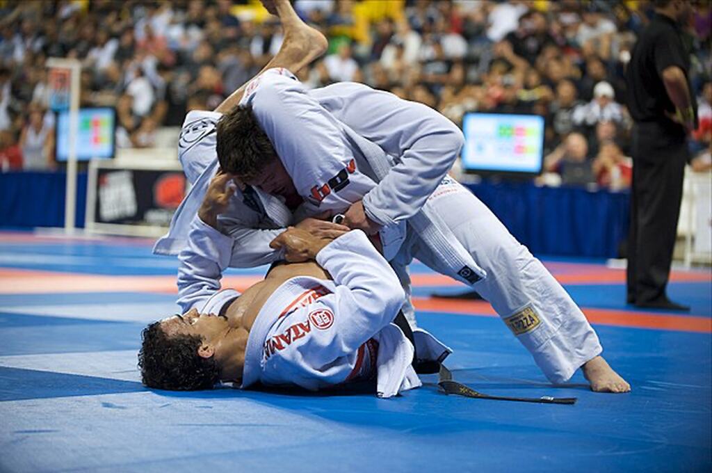 Dois lutadores de Jiu-Jitsu vestindo kimonos brancos, lutando em tatame azul, com prendendo a cabeça do outro em suas pernas.