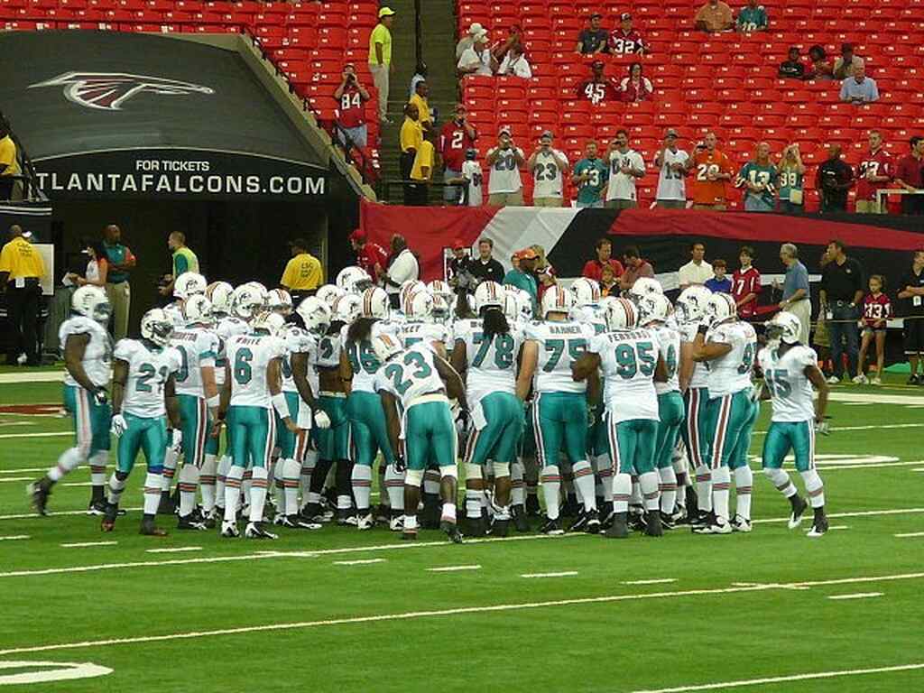 Equipe dos Miami Dolphins, com seu uniforme branco e verde, reunida e abraçada em campo diante da torcida nas arquibancadas.