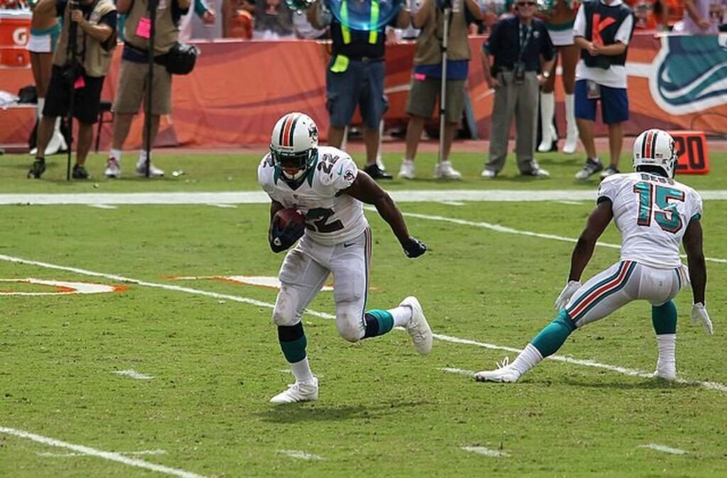 Jogador dos Miami Dolphins de uniforme branco, correndo com a bola ao lado de seu companheiro de equipe, durante a partida.