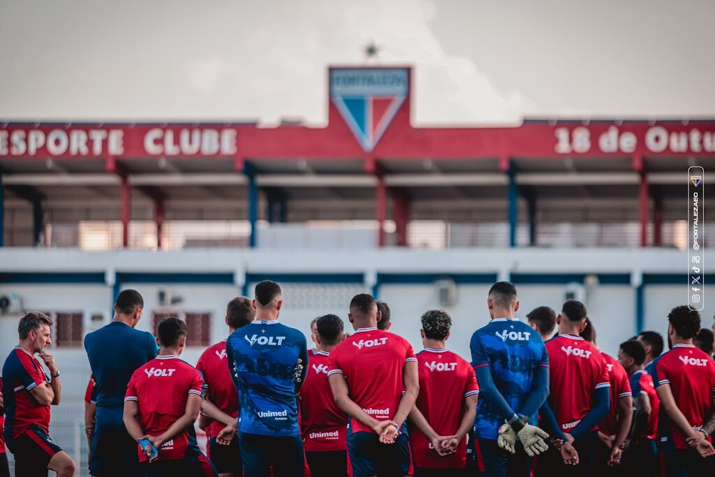 Jogadores e comissão técnica do Fortaleza, vestindo uniformes vermelhos e azuis, em frente a arquibancada com escudo do time.