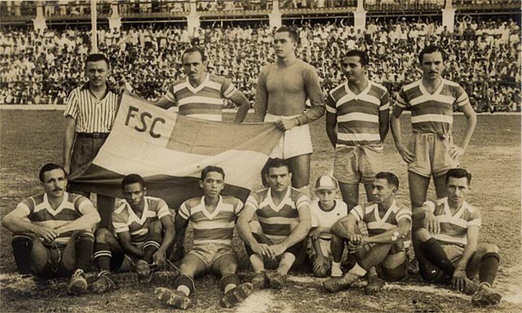 Time antigo do Fortaleza, com jogadores enfileirados segurando bandeira do clube no gramado, antes da partida.