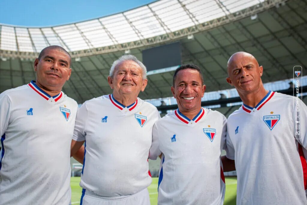 Ex-jogadores do Fortaleza, já aposentados, vestindo novo uniforme branco do time e abraçados no estádio.