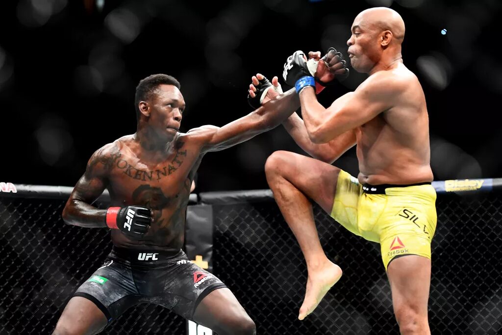 Lutador de UFC Anderson Silva, vestindo calção amarelo, dando chute em direção ao Israel Adesanya.