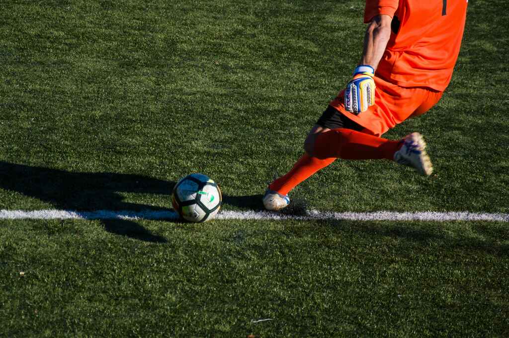 Goleiro de futebol, vestindo uniforme laranja, chutando a bola em cobrança de tiro de meta.