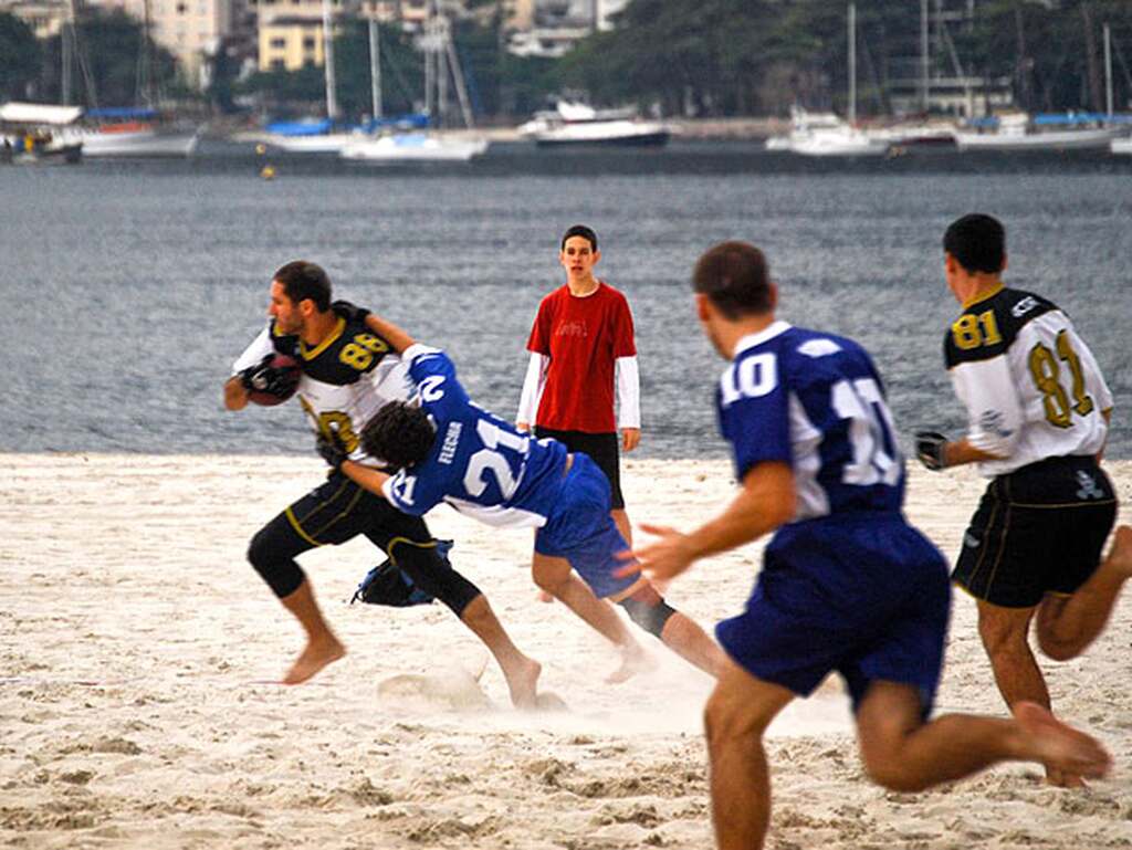 Homens jogando Carioca Bowl na praia, precursor do futebol americano no Brasil.