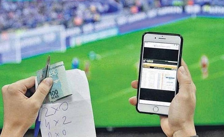 Apostador iniciante usa um celular para fazer apostas esportivas. Ao fundo, um estádio de futebol