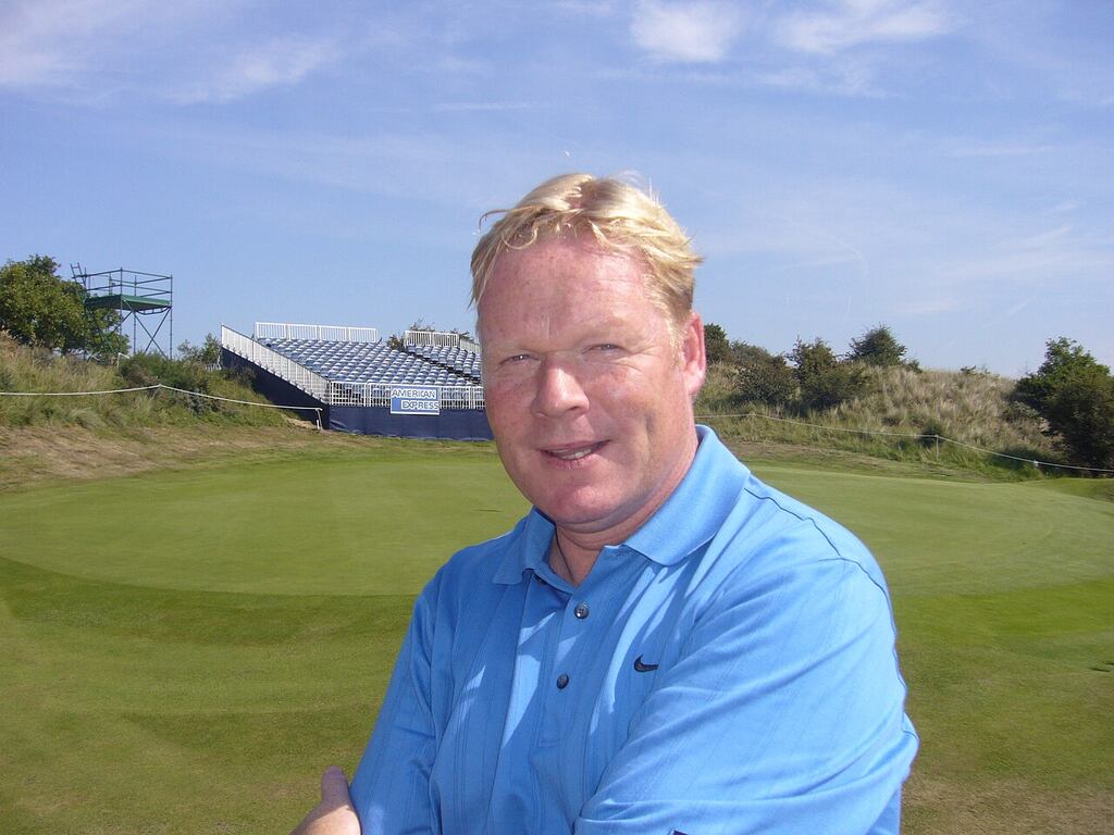 Imagem do ex zagueiro Ronald Koeman sorrindo, com um gramado e uma arquibancada ao fundo