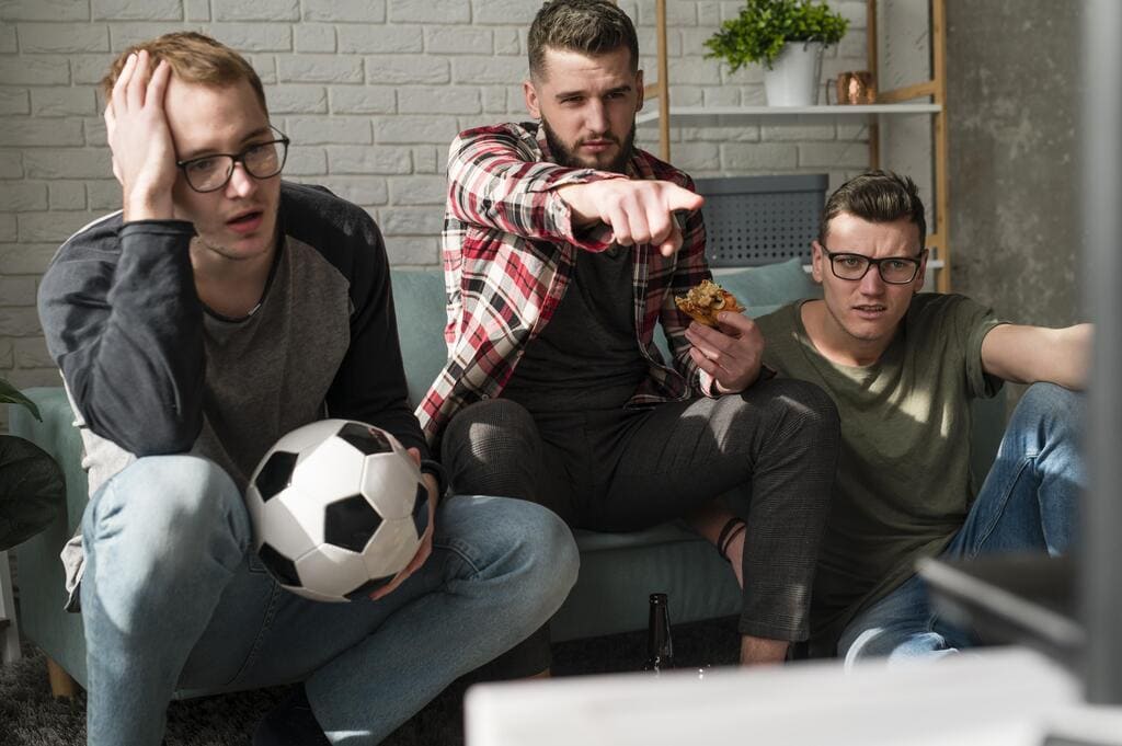 3 homens sentados em sofá, segurando uma bola de futebol e apontando para frente com atenção.