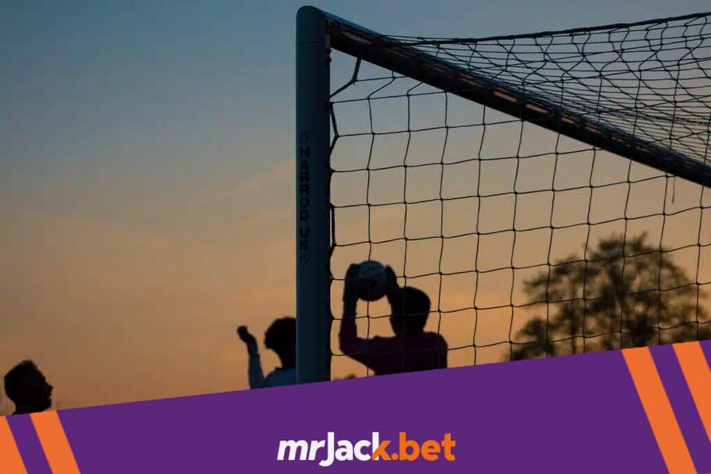 Meninos jogando futebol ao ar livre ao anoitecer, com o goleiro agarrando a bola.