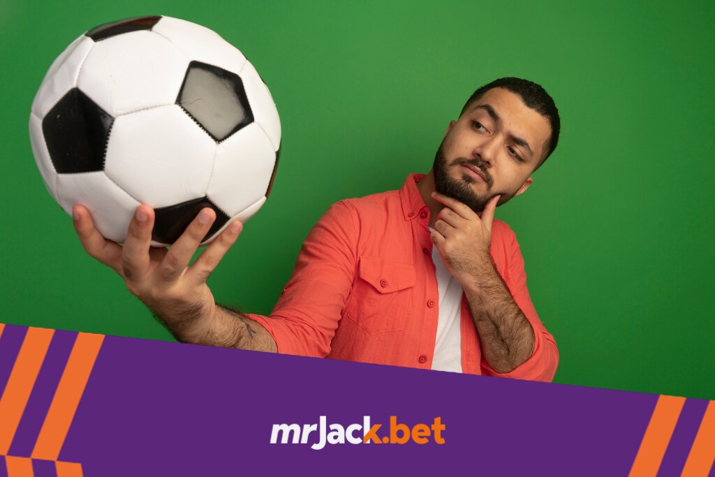 Homem vestindo camisa branca e blusa laranja, em um fundo verde, segurando uma bola de futebol e pensando.