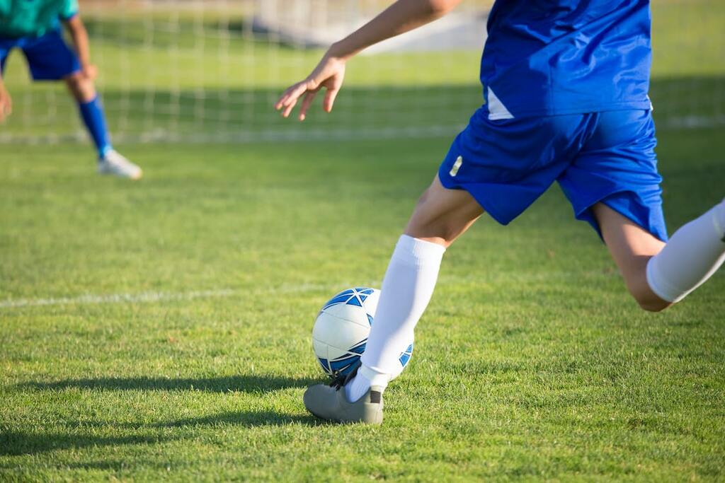 Jogador de futebol vestindo uniforme azul com detalhes brancos, realizando chute com a perna direita em direção ao gol.