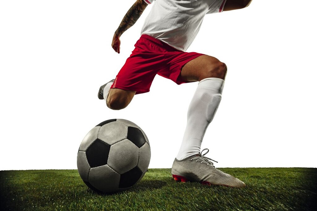 Jogador de futebol de camisa branca e bermuda vermelha chutando bola de futebol clássica com o pé direito.