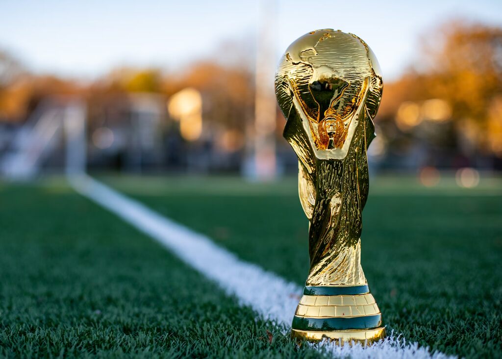 Taça da Copa do Mundo colocada sobre a linha de fundo de um campo de futebol.