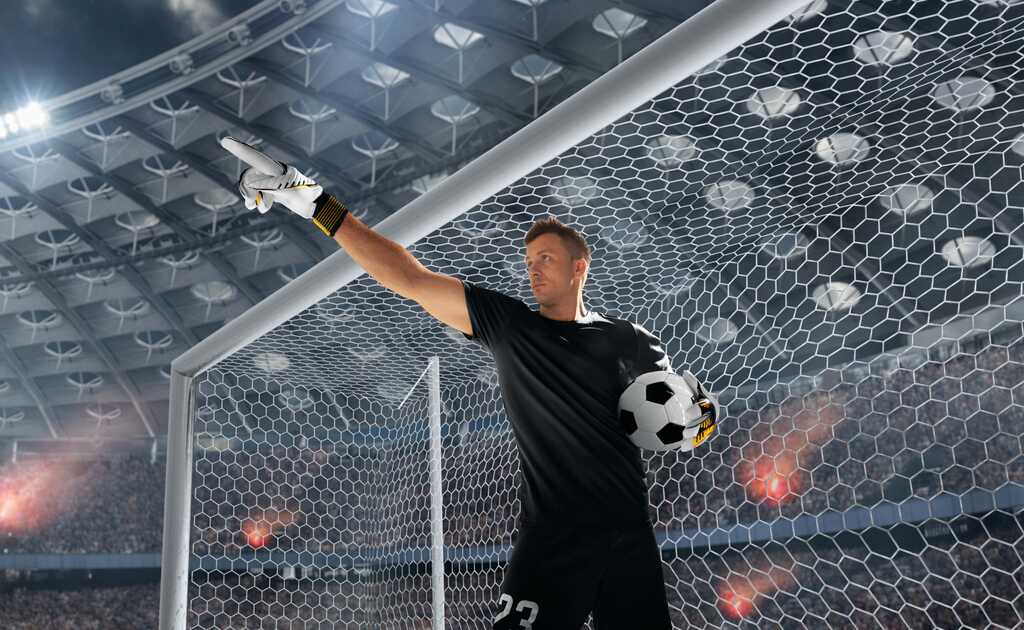 Goleiro de futebol segurando uma bola debaixo de um dos braços apontando para frente.