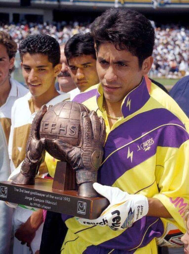 Ex-jogador de futebol Jorge Campos segurando uma premiação para goleiros, enquanto veste seu uniforme amarelo.