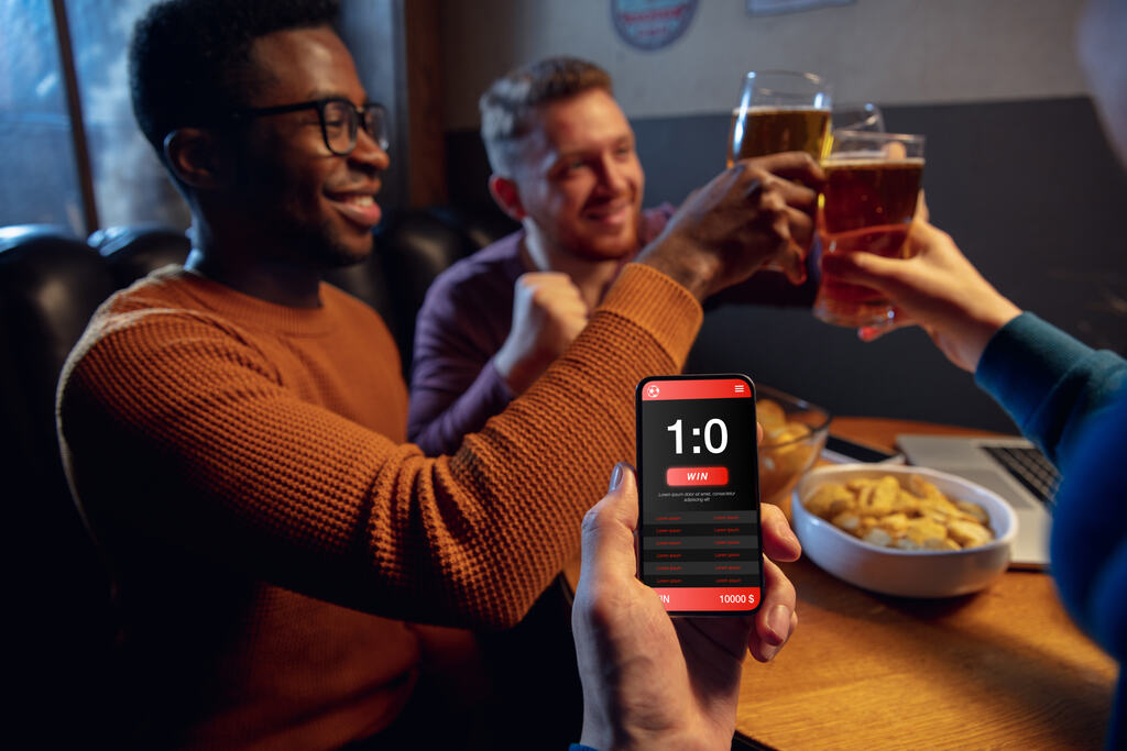 Homens tomando cerveja em um bar enquanto olham resultado de aposta esportiva em aplicativo no celular.