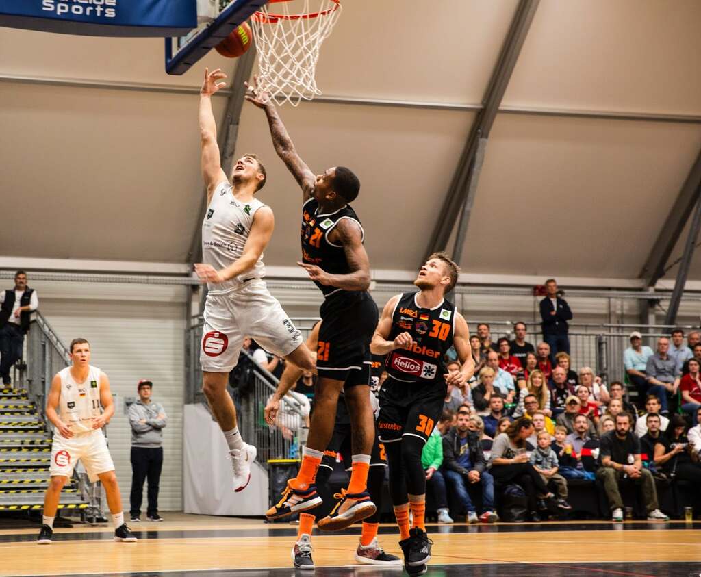 Jogador de basquete vestindo uniforme branco tentando realizar uma bandeja durante o jogo, enquanto jogador de preto bloqueia.