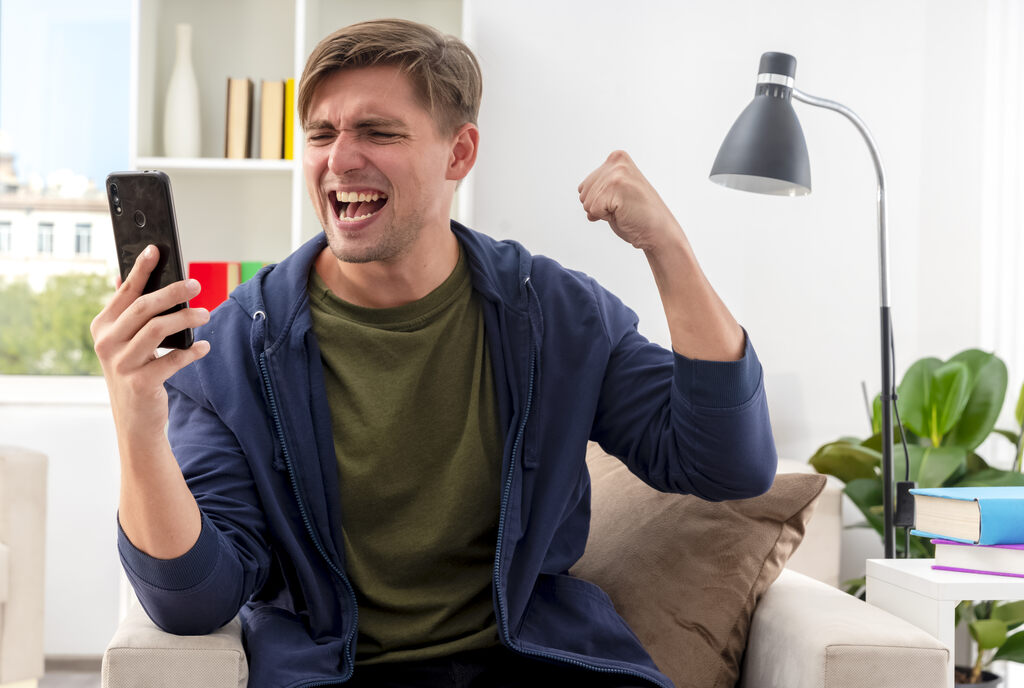 Homem de camisa verde e casaco azul comemorando algo enquanto segura um celular com a mão direita.