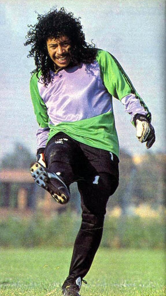 Ex-jogador de futebol René Higuita, vestindo uniforme verde de goleiro com a perna direita levantada.