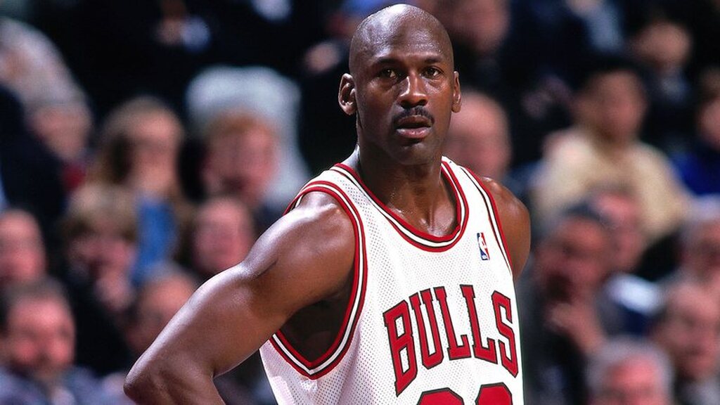 Jogador de basquete Michael Jordan, jogando durante partida com uniforme branco, com as mãos na cintura.