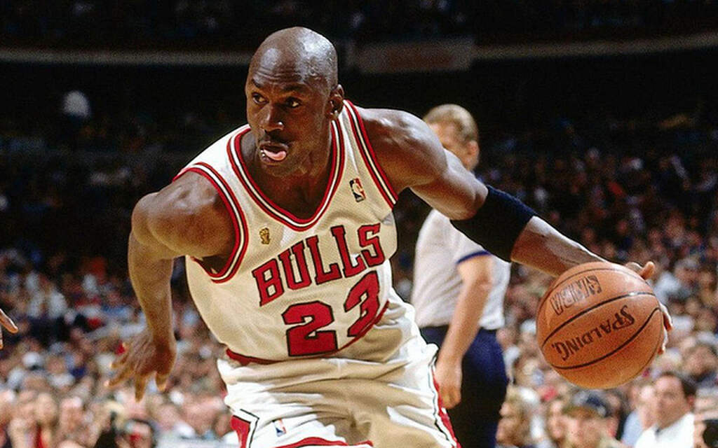 Jogador de basquete Michael Jordan, de uniforme branco conduzindo a bola durante a partida.