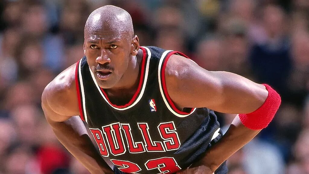 Jogador de basquete Michael Jordan, vestindo uniforme preto, agachado, durante partida de basquete.