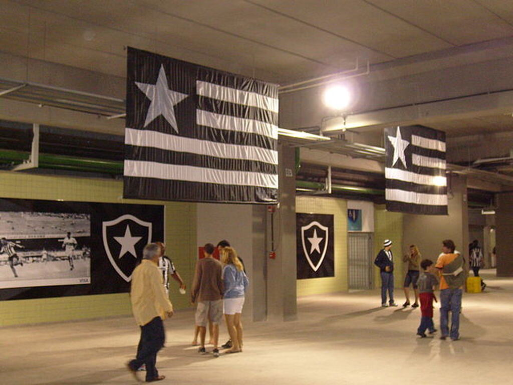 Salão com diversas pessoas andando de um lado para o outro, cheio de bandeiras do Botafogo espalhadas.