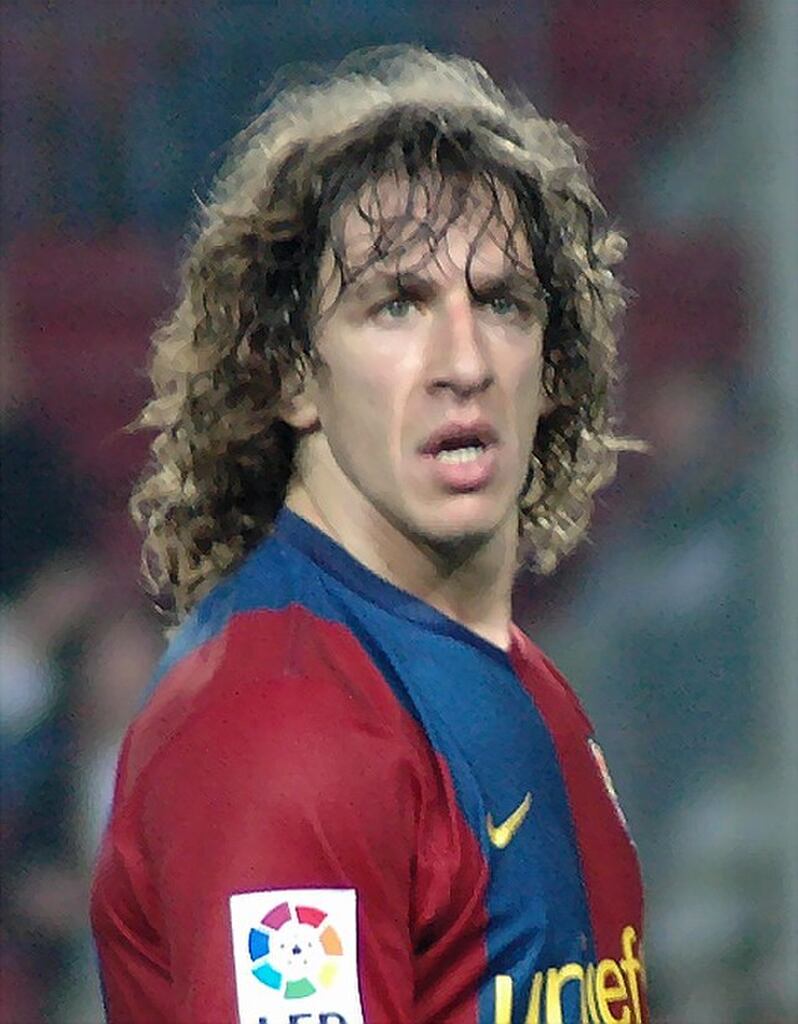 Ex-jogador de futebol do Barcelona, Puyol, usando uniforme vermelho e azul da equipe, e andando pelo campo durante partida.