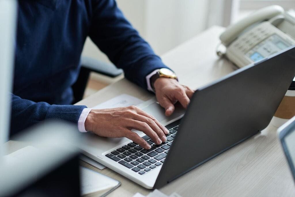 Homem de blusa azul, com relógio no pulso esquerda, digitando em um notebook sobre mesa de madeira.