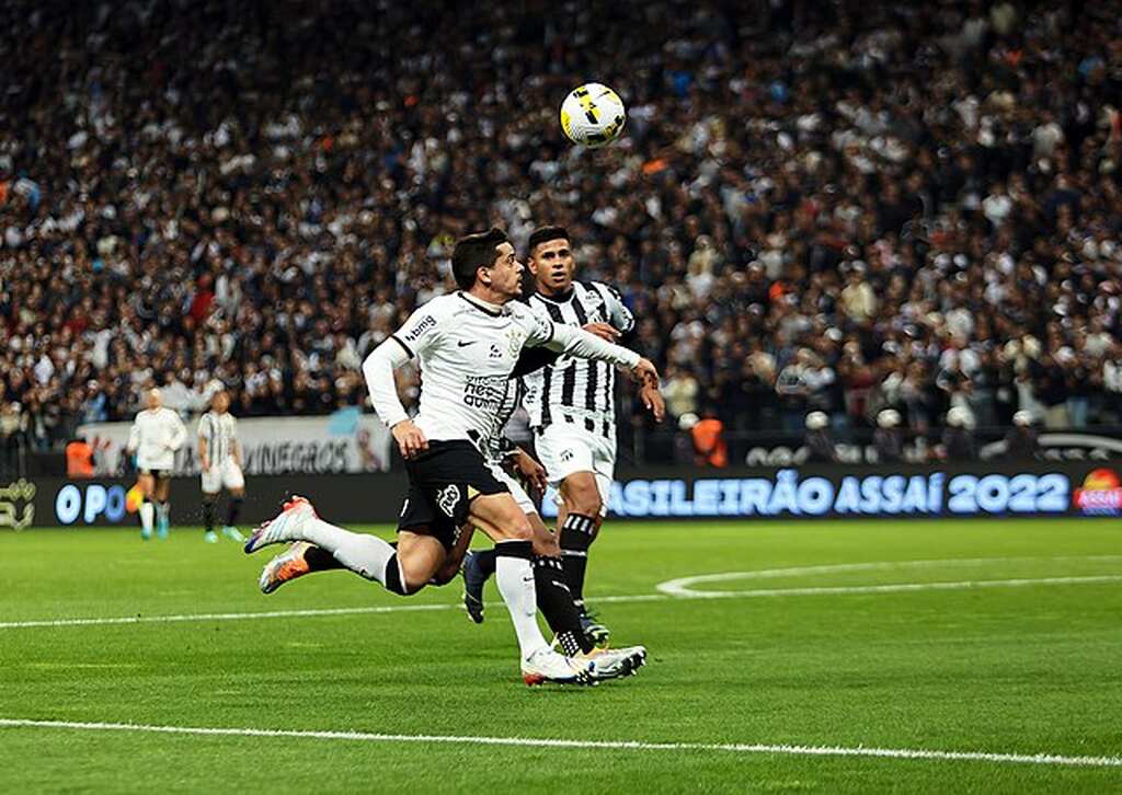 Jogador de futebol do Corinthians disputando uma bola dentro da área com jogador adversário.