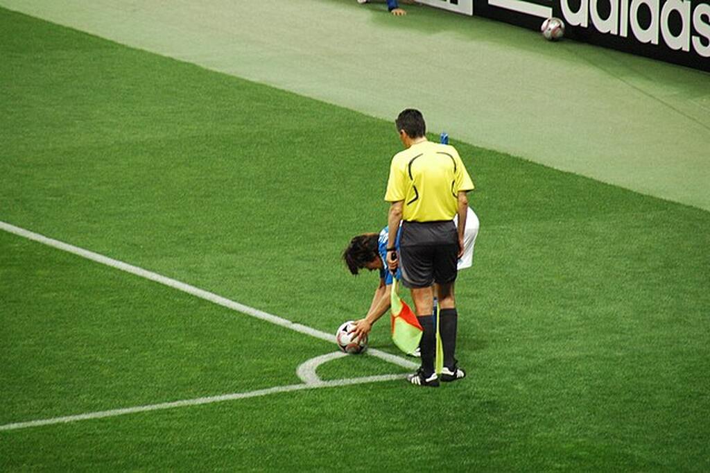 Jogador de uniforme azul, arrumando a bola para cobrança de escanteio ao lado do bandeirinha.