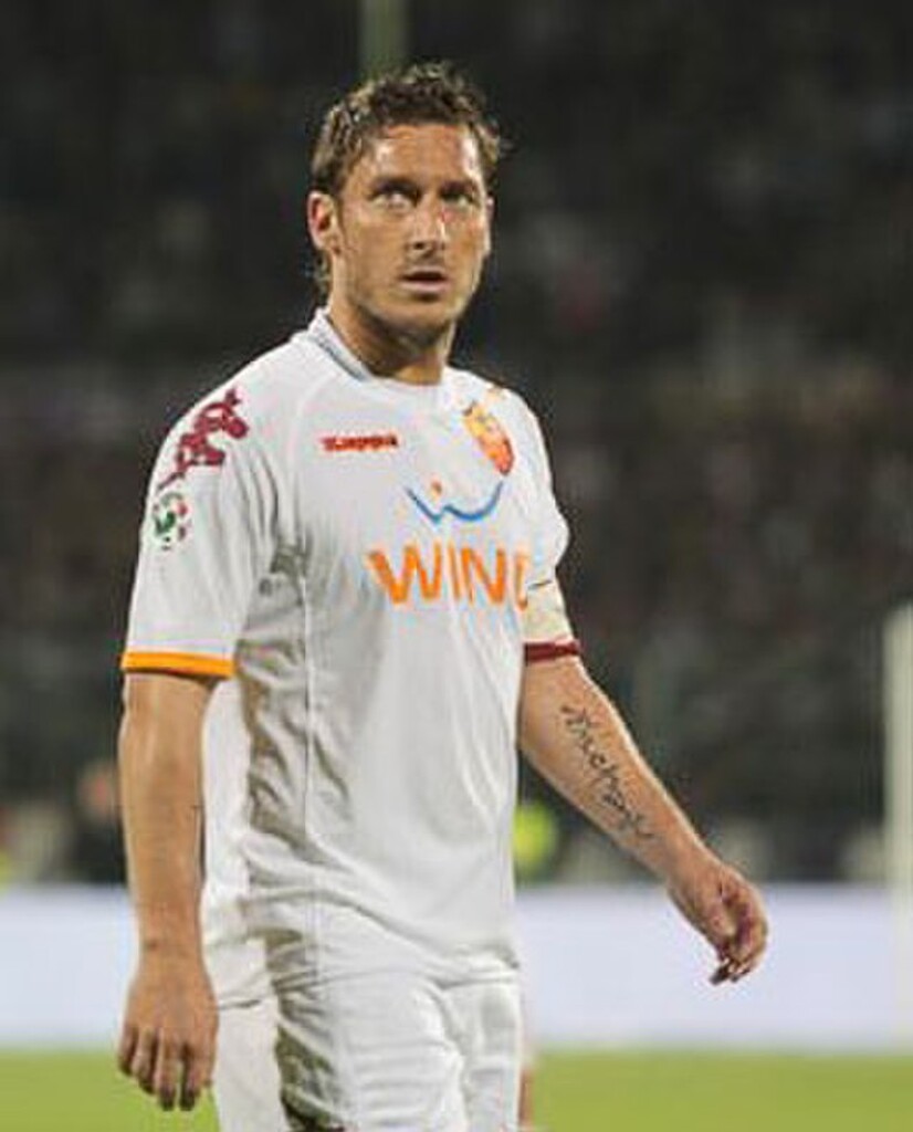 Ex-jogador de futebol Francesco Totti, vestindo uniforme branco da Roma, e andando pelo campo durante partida de futebol.