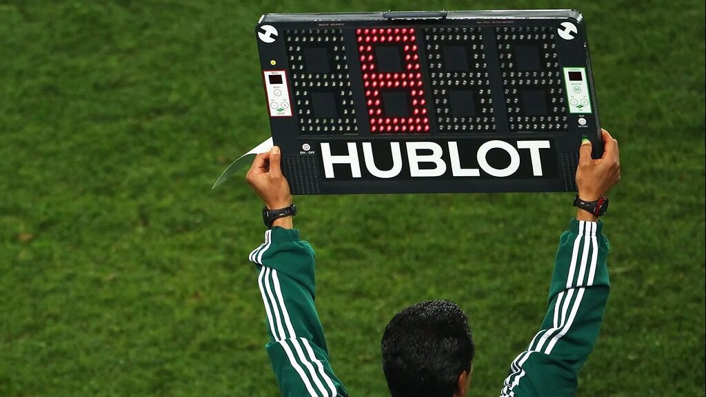 Árbitro auxiliar, vestindo uniforme verde, erguendo a placa com os minutos adicionais do jogo.