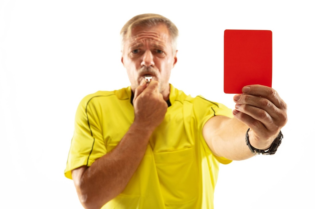 Árbitro de futebol com uniforme amarelo, apito na boca, e mostrando o cartão vermelho.