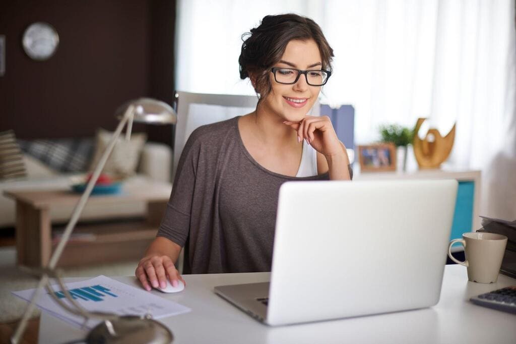 Mulher de roupa cinza, de óculos, usando um notebook sobre uma mesa branca, sorrindo.