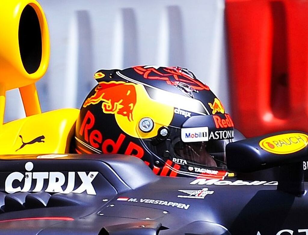 Piloto de Fórmula 1 usando capacete preto com detalhes vermelhos e amarelos da Red Bull, dentro de carro do carro durante corrida.