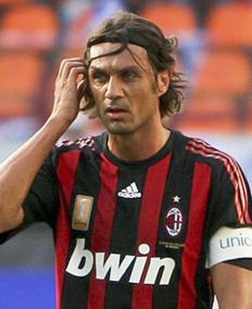 Ex-jogador de futebol Paolo Maldini, vestindo camisa vermelha e preta do Milan, usando faixa de capitão, durante partida de futebol.