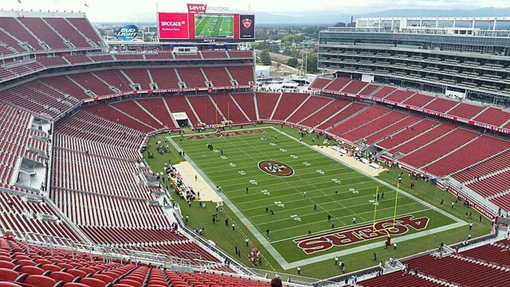 Estádio de futebol americano visto do alto, com arquibancadas com cadeiras vermelhas vazias, e pessoas andando pelo campo.