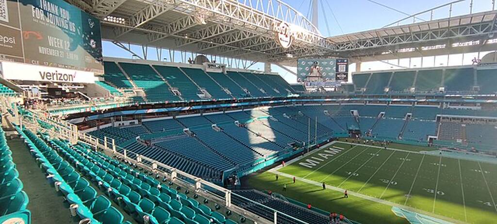 Estádio de futebol americano, com vista da arquibancada para as cadeiras azuis vazias, em um dia de sol.