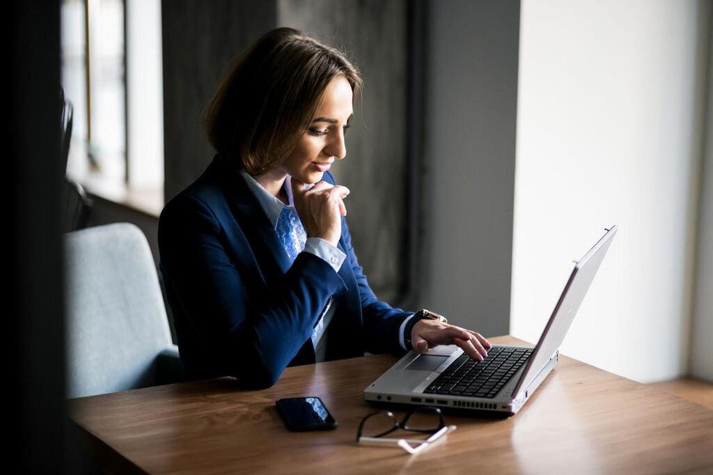 Mulher de roupa social, utilizando um notebook com a mão esquerda enquanto encosta a direita no queixo pensativa.