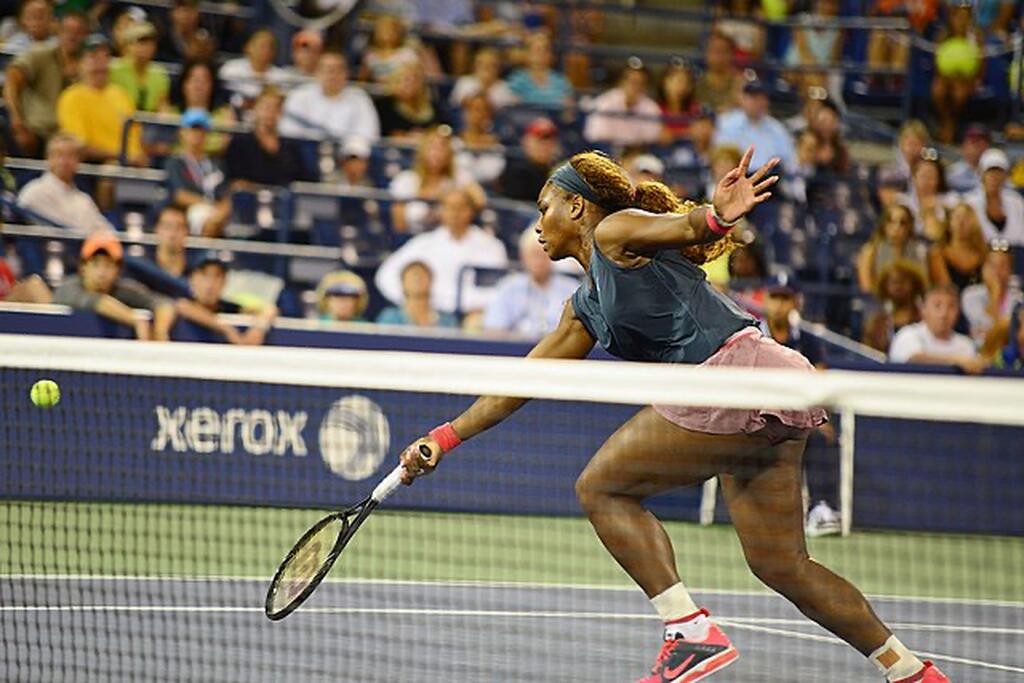 Ex-jogadora de tênis, Serena Williams, vestindo camisa verde-escura, e short rosa, correndo para rebater a bola durante a partida.