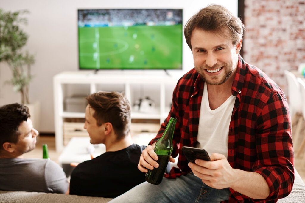 Homem segurando um smartphone e uma garrafa de cerveja, enquanto outros dois conversam sentados no sofá e um jogo passa na TV