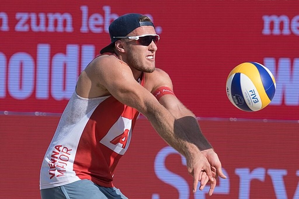 Jogador de vôlei de praia, de regata vermelha e branca, boné e óculos escuro, durante partida, recebendo a bola de manchete.