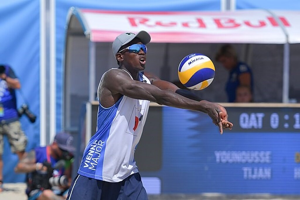 Jogador de vôlei de praia, de regata branca e azul, boné e óculos escuro, durante partida, recebendo a bola de manchete.