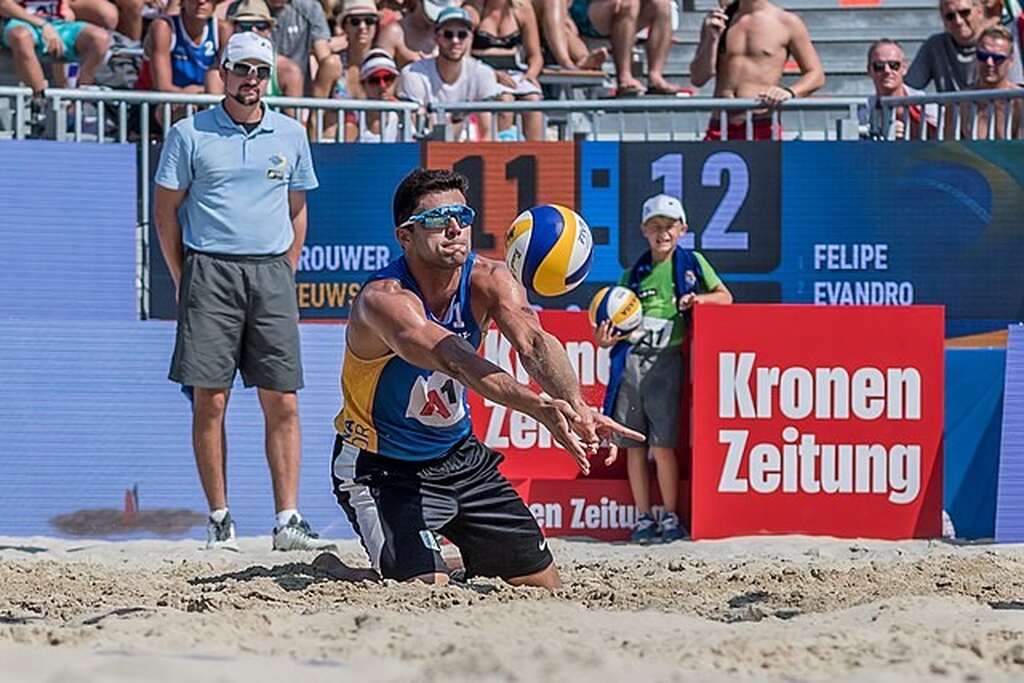 Jogador de vôlei de praia, de regata azul e amarela, calção preto e óculos escuro, recebendo a bola de manchete.