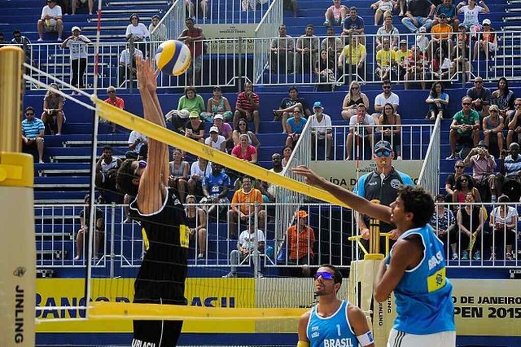 Jogador de vôlei de praia bloqueando bola cortada por jogador adversário, do outro lado da rede, durante a partida.