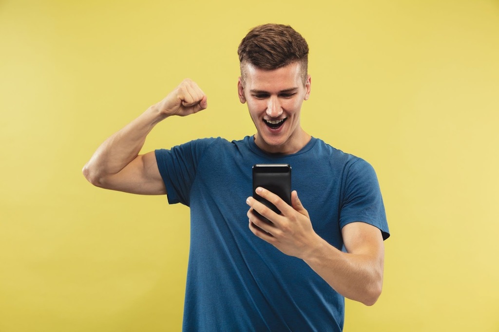 Homem de camiseta azul sorrindo e comemorando, enquanto segura e olha para um smartphone diante de um fundo amarelo.