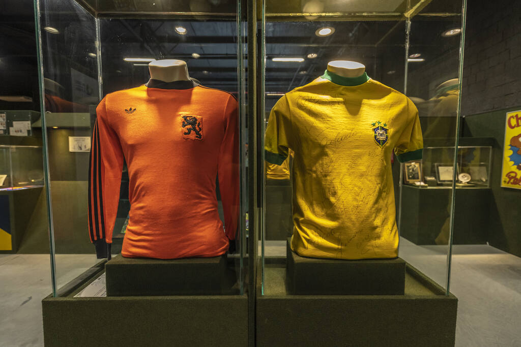 Camisa histórica da seleção holandesa ao lado de camisa da seleção brasileira, ambas em um museu.