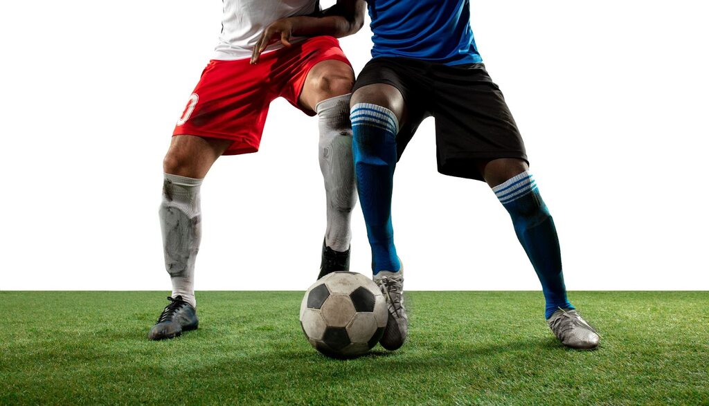 Jogador de futebol de camisa branca e cação vermelho, disputando bola com jogador de uniforme azul e calção preto.